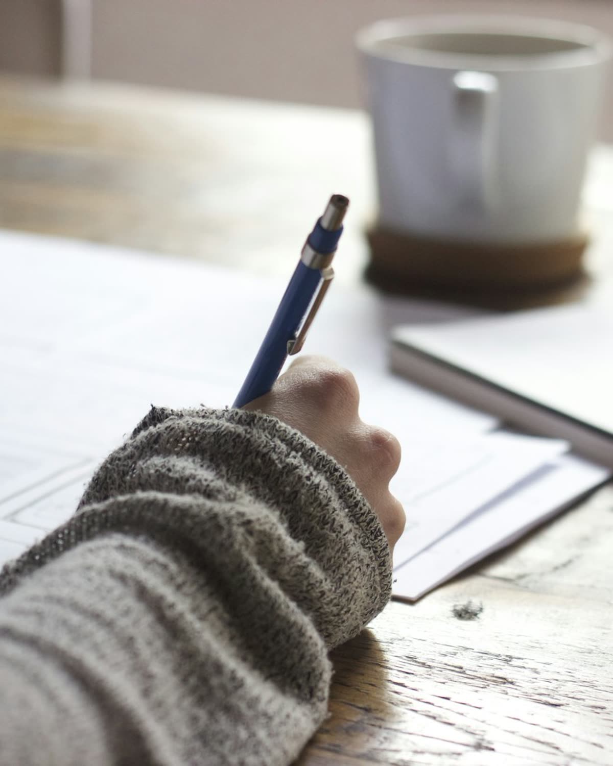 Hands writing in a notebook on a wooden desk beside loose papers and a pen.